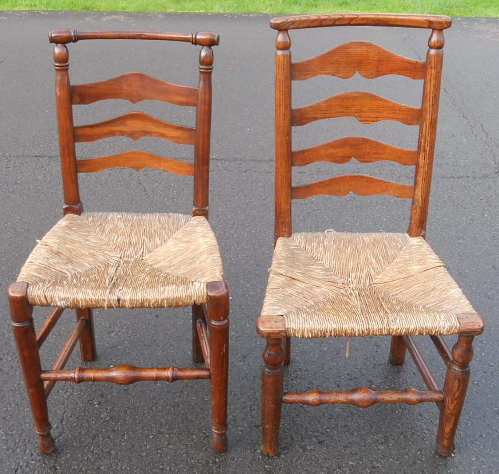 A pair of antique elm dining chairs featuring shaped ladderback rails, woven rush seats, and turned wooden legs.