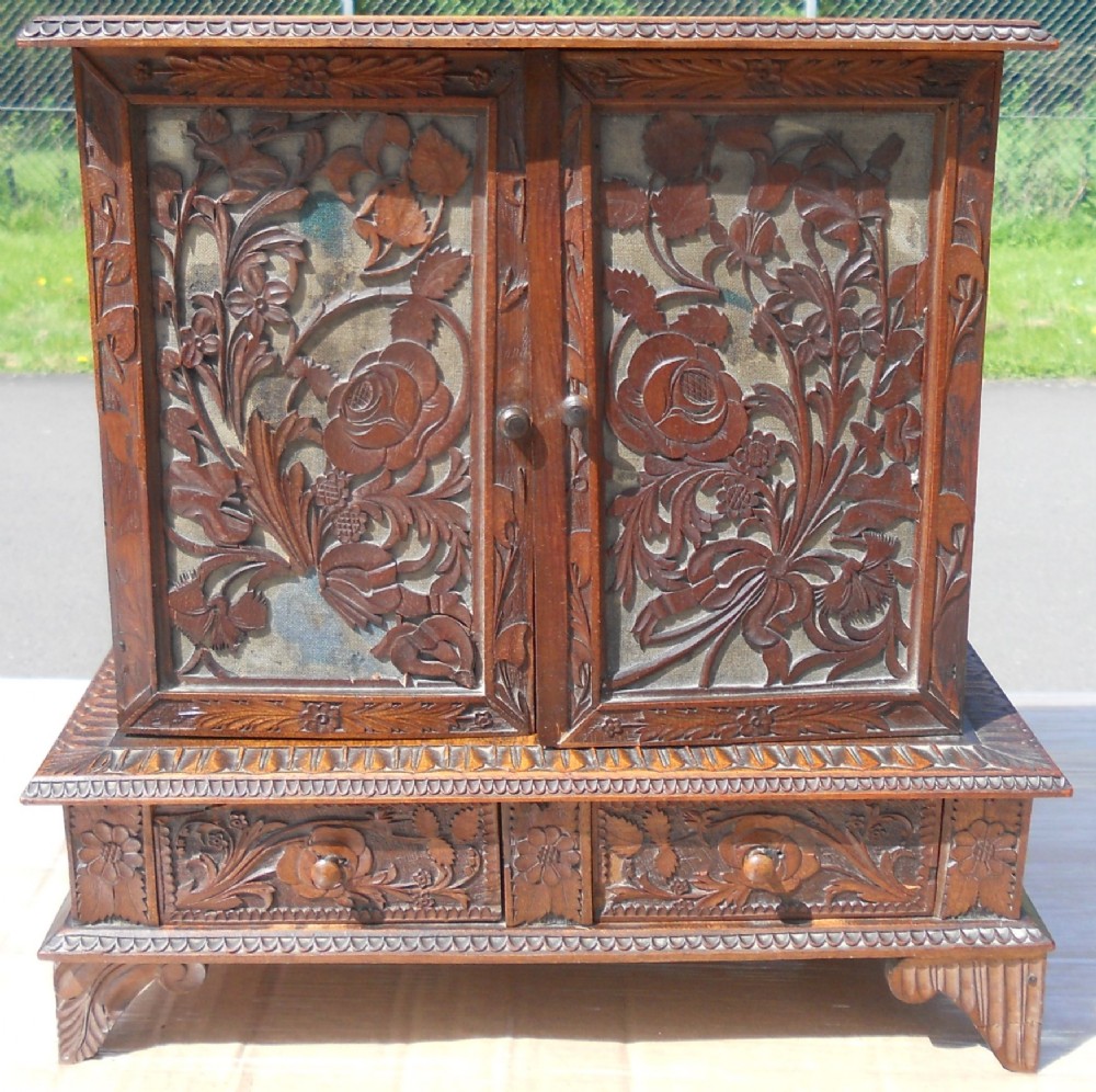 A small dark brown carved wooden tabletop cabinet with two doors featuring openwork floral designs backed by fabric and two lower carved drawers.
