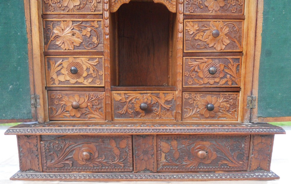 A small dark brown carved wooden tabletop cabinet with two doors featuring openwork floral designs backed by fabric and two lower carved drawers. View VII