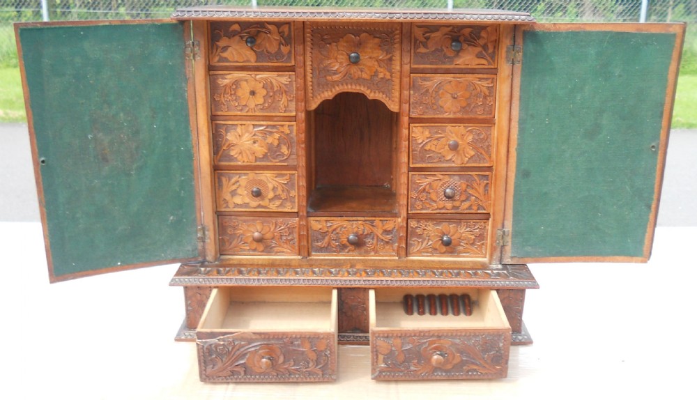 A small dark brown carved wooden tabletop cabinet with two doors featuring openwork floral designs backed by fabric and two lower carved drawers. View VIII