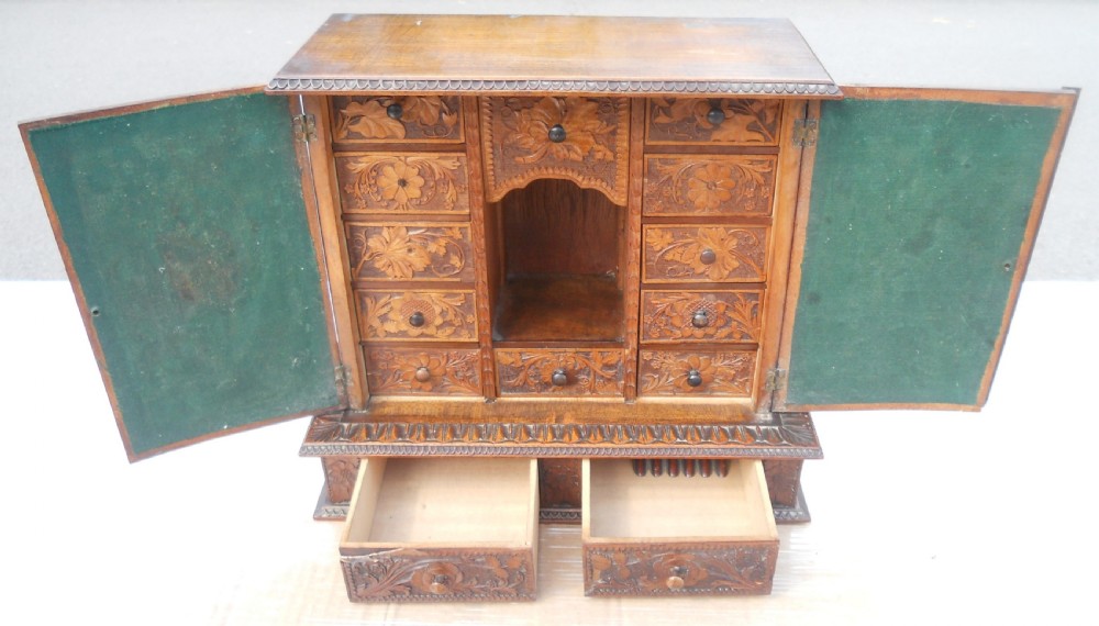 A small dark brown carved wooden tabletop cabinet with two doors featuring openwork floral designs backed by fabric and two lower carved drawers. View IX