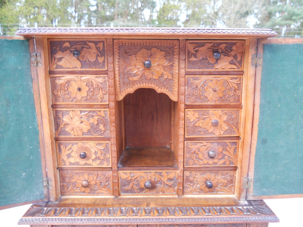A small dark brown carved wooden tabletop cabinet with two doors featuring openwork floral designs backed by fabric and two lower carved drawers. View X