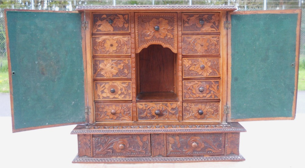 A small dark brown carved wooden tabletop cabinet with two doors featuring openwork floral designs backed by fabric and two lower carved drawers. View XI
