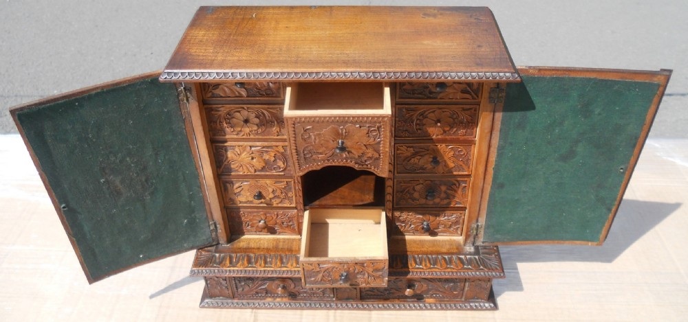 A small dark brown carved wooden tabletop cabinet with two doors featuring openwork floral designs backed by fabric and two lower carved drawers. View XIV