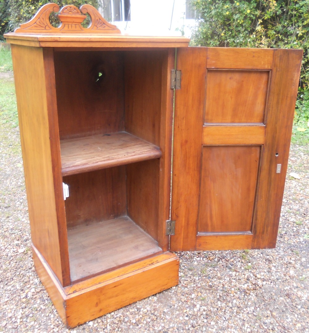 An Edwardian satin walnut bedside cabinet featuring a carved scroll cresting rail, a panelled door with relief carving, and a brass drop handle resting on a solid plinth base. View VI