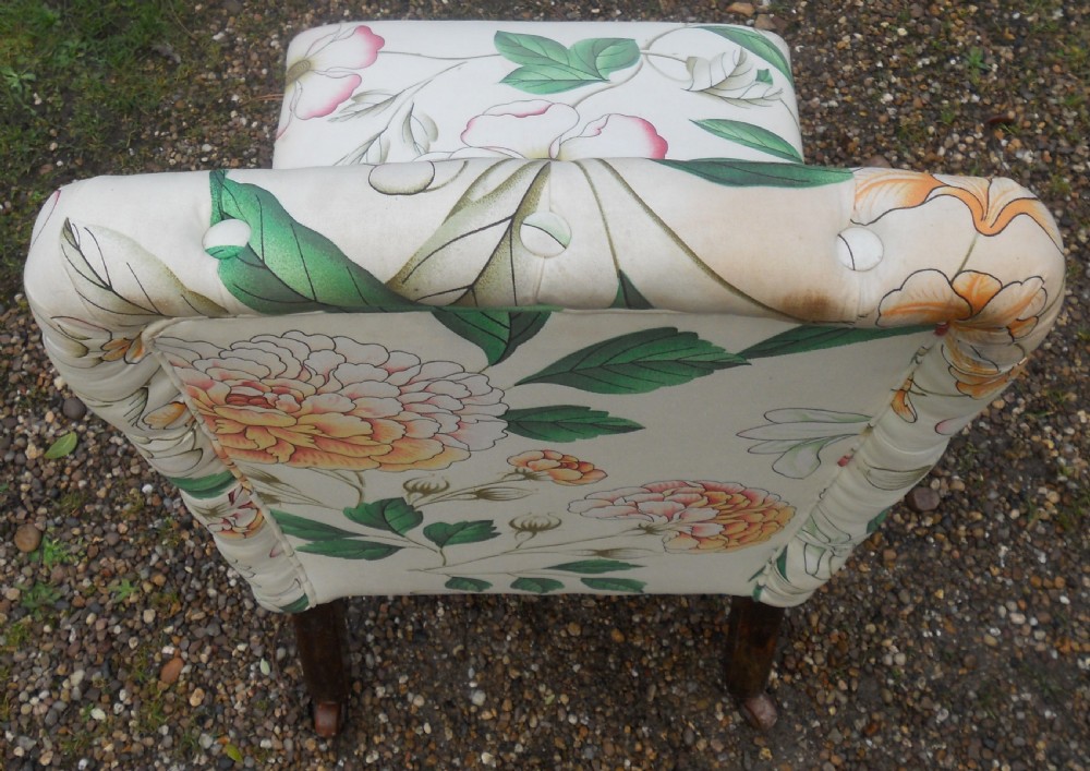 An Edwardian low-seated nursing chair featuring floral patterned upholstery, deep buttoning on the backrest, braided trim, and dark turned wooden front legs on castors. View V