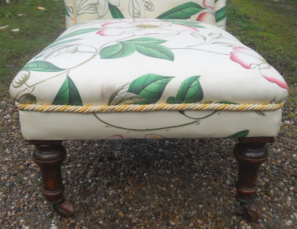 An Edwardian low-seated nursing chair featuring floral patterned upholstery, deep buttoning on the backrest, braided trim, and dark turned wooden front legs on castors. View VI