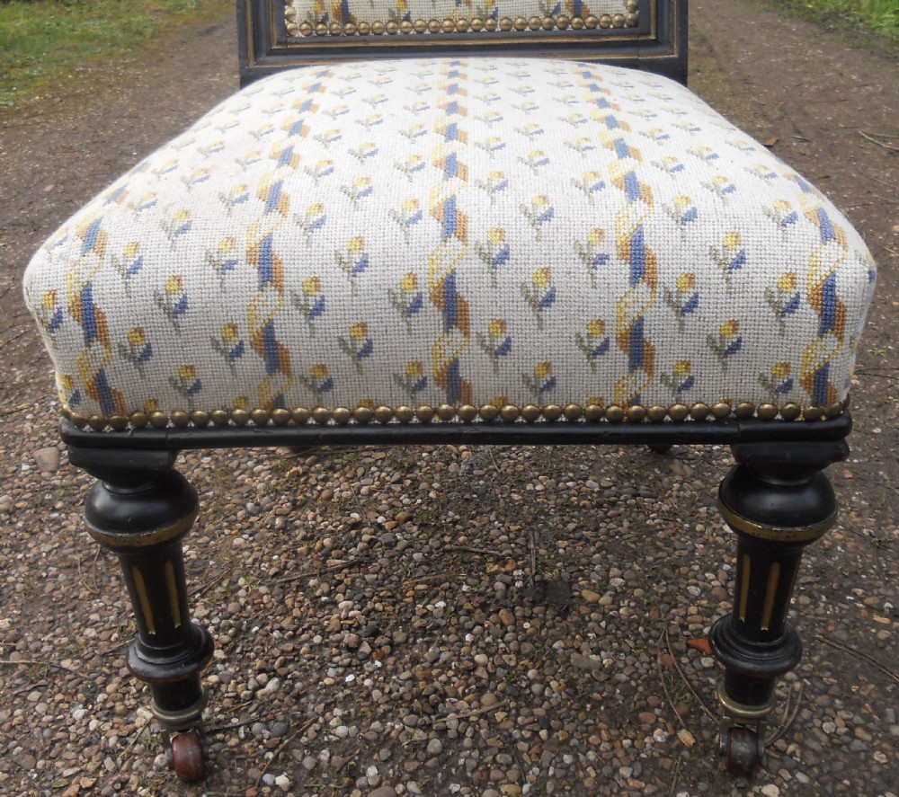 An ebonised Victorian nursing chair featuring cream floral needlepoint upholstery, brass studding, turned front legs on castors, and gilt linear detailing. View IV