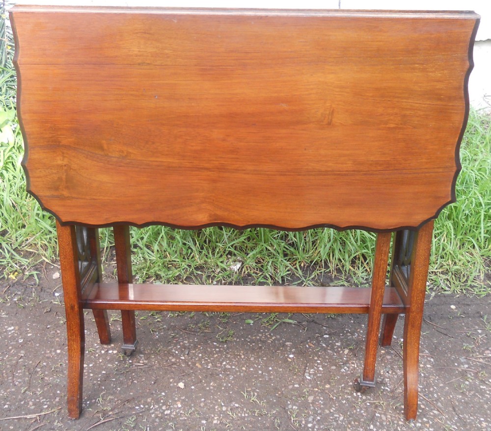 An Edwardian mahogany drop-leaf occasional table featuring scalloped edges, a lower stretcher shelf, and slender legs on brass castors.
