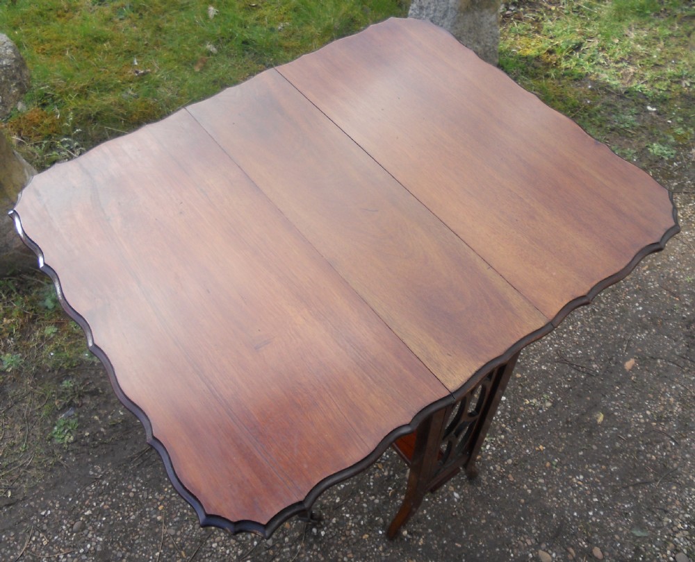 An Edwardian mahogany drop-leaf occasional table featuring scalloped edges, a lower stretcher shelf, and slender legs on brass castors. View VIII