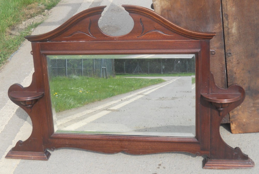 An Edwardian mahogany overmantel mirror featuring a shaped pediment with incised carvings, twin side display shelves, and a bevelled mirror plate.