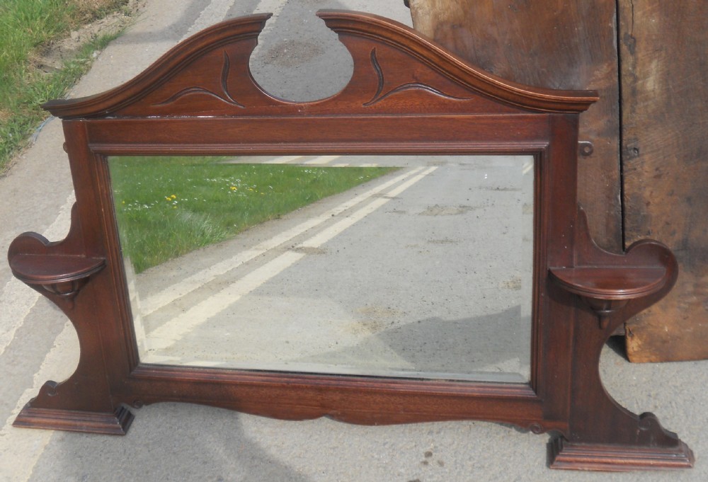 An Edwardian mahogany overmantel mirror featuring a shaped pediment with incised carvings, twin side display shelves, and a bevelled mirror plate. View II