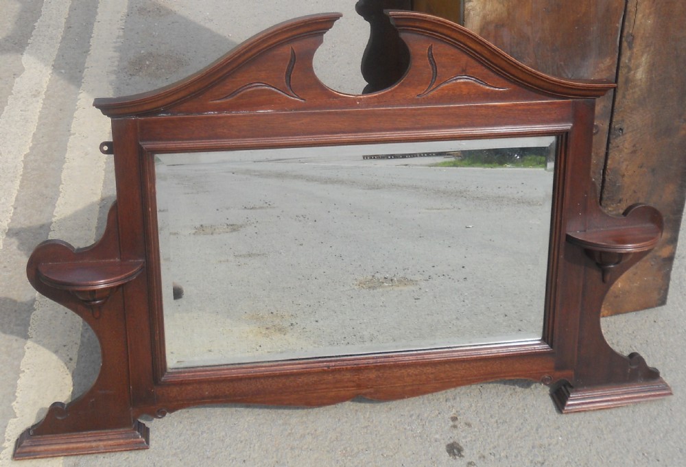 An Edwardian mahogany overmantel mirror featuring a shaped pediment with incised carvings, twin side display shelves, and a bevelled mirror plate. View III