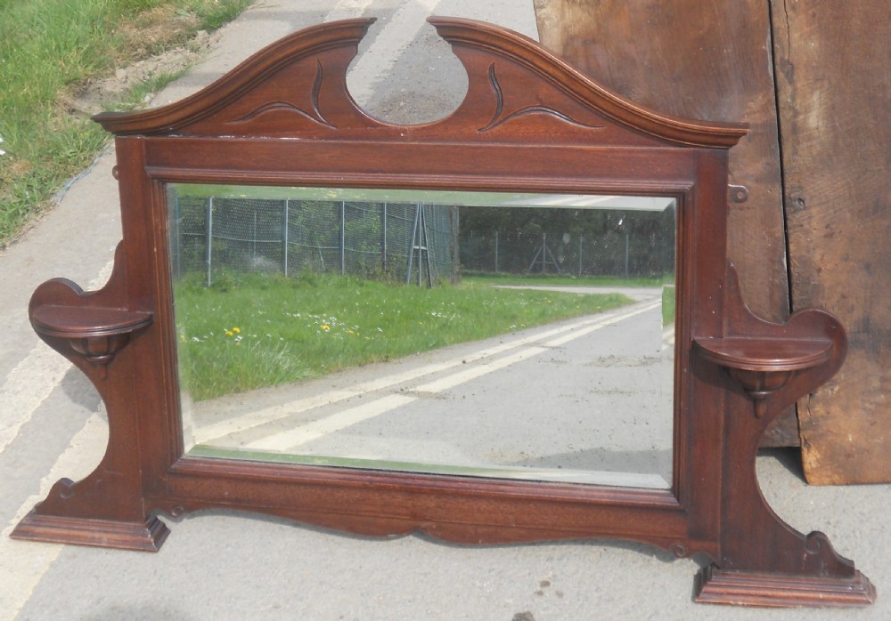 An Edwardian mahogany overmantel mirror featuring a shaped pediment with incised carvings, twin side display shelves, and a bevelled mirror plate. View IV