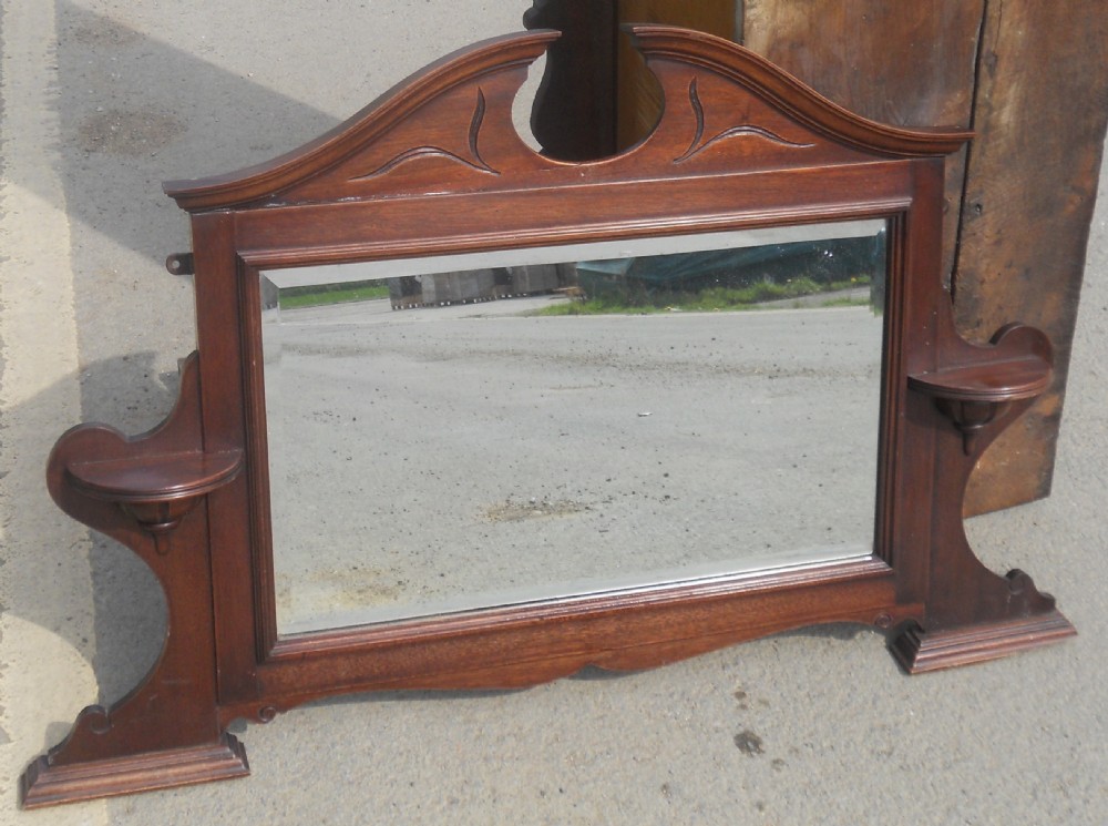 An Edwardian mahogany overmantel mirror featuring a shaped pediment with incised carvings, twin side display shelves, and a bevelled mirror plate. View V