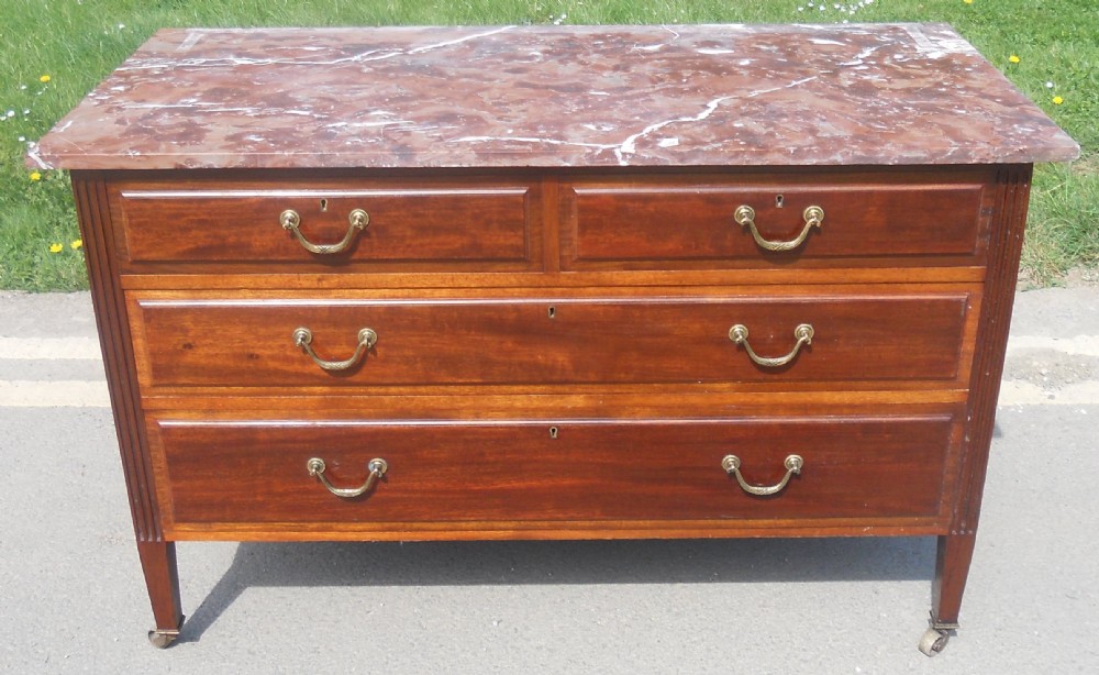 An Edwardian mahogany chest of drawers featuring a variegated red marble top, brass drop handles, and tapered legs on castors.