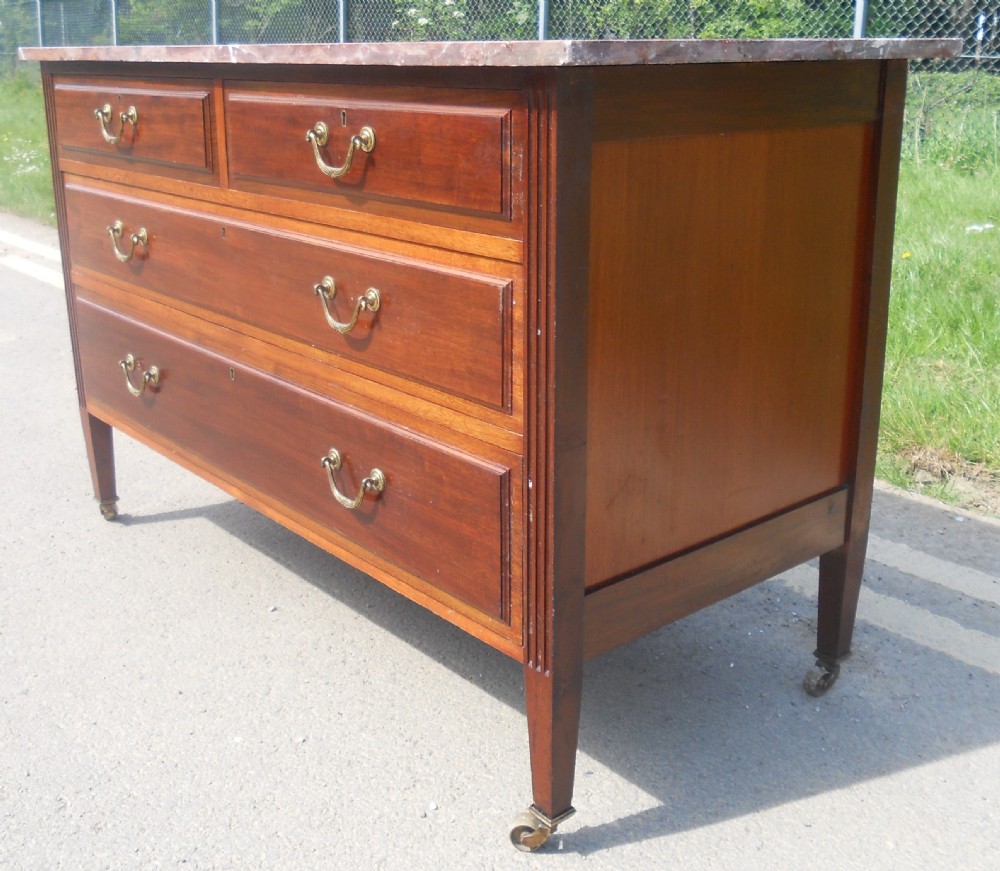 An Edwardian mahogany chest of drawers featuring a variegated red marble top, brass drop handles, and tapered legs on castors. View II