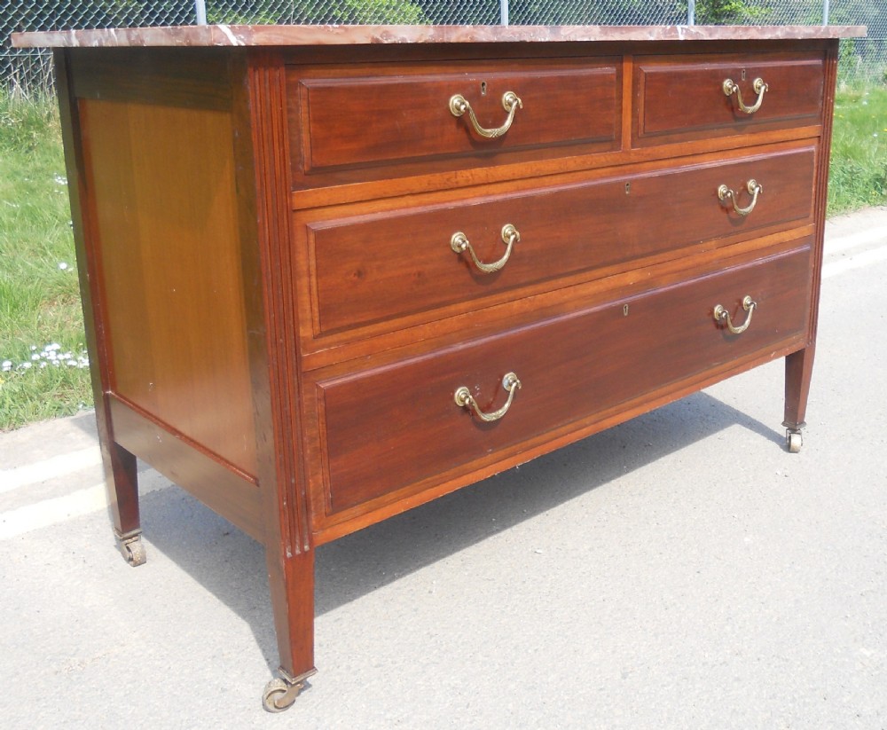 An Edwardian mahogany chest of drawers featuring a variegated red marble top, brass drop handles, and tapered legs on castors. View III
