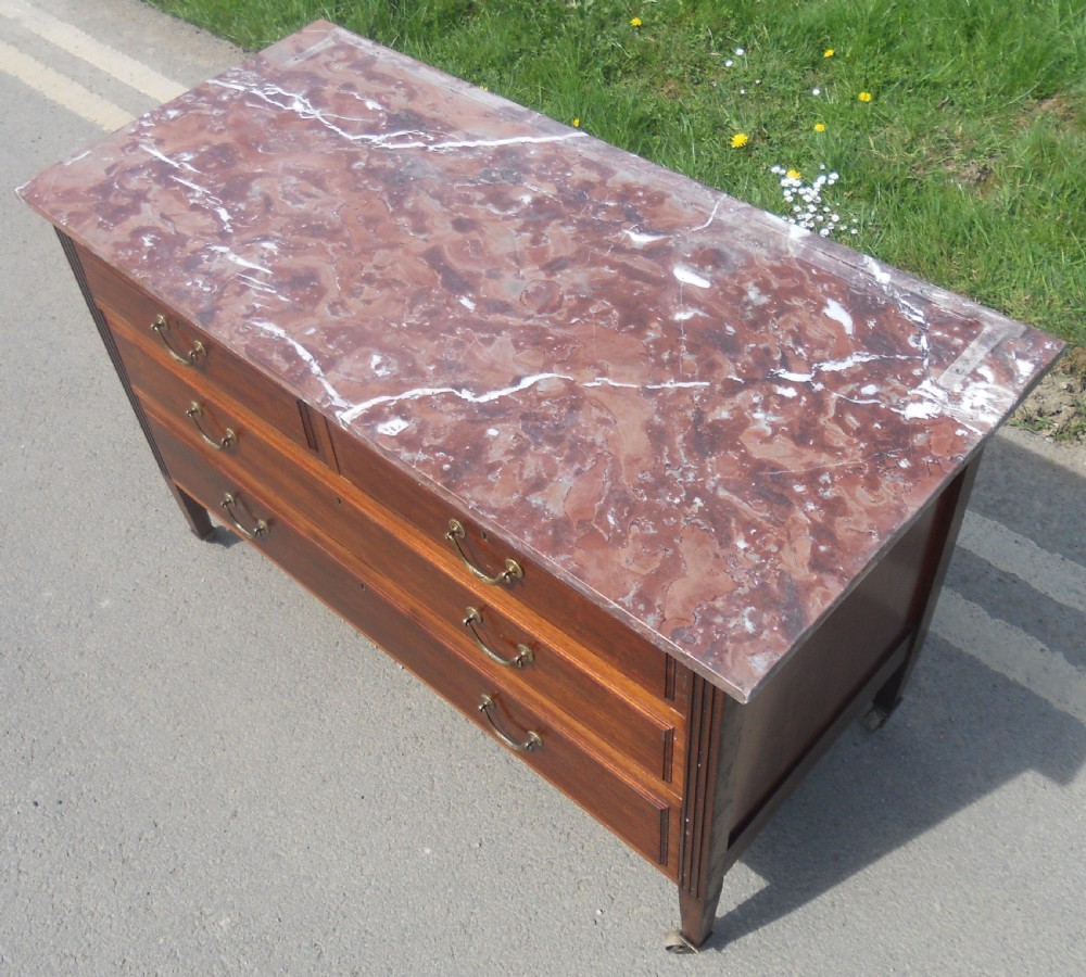 An Edwardian mahogany chest of drawers featuring a variegated red marble top, brass drop handles, and tapered legs on castors. View V