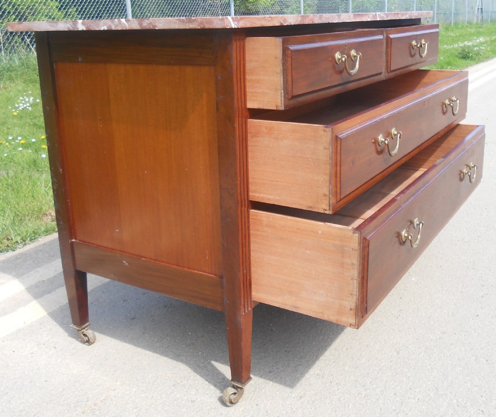 An Edwardian mahogany chest of drawers featuring a variegated red marble top, brass drop handles, and tapered legs on castors. View IX
