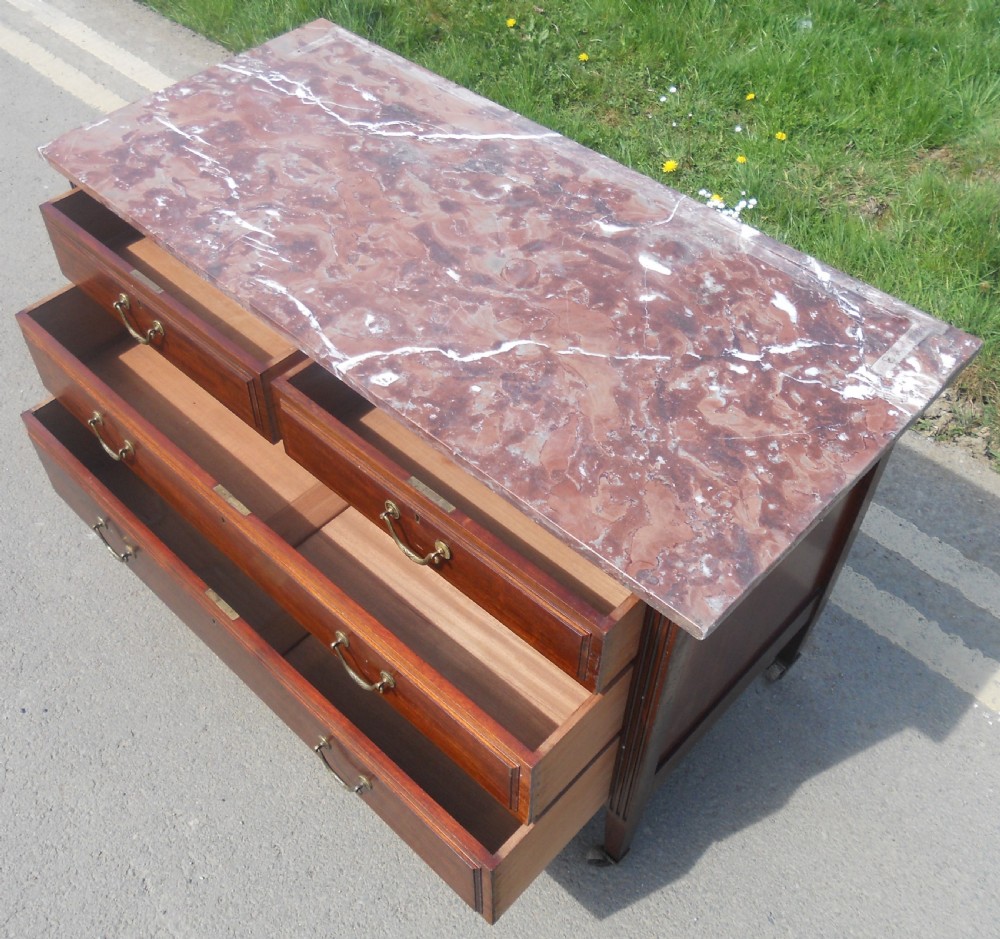 An Edwardian mahogany chest of drawers featuring a variegated red marble top, brass drop handles, and tapered legs on castors. View X