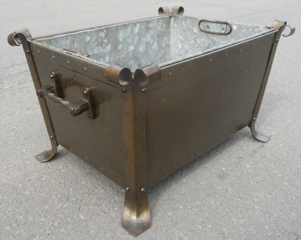 A rectangular patinated metal log bin featuring riveted borders, scrolled corner finials, side handles, splayed feet, and a fitted galvanised metal liner.