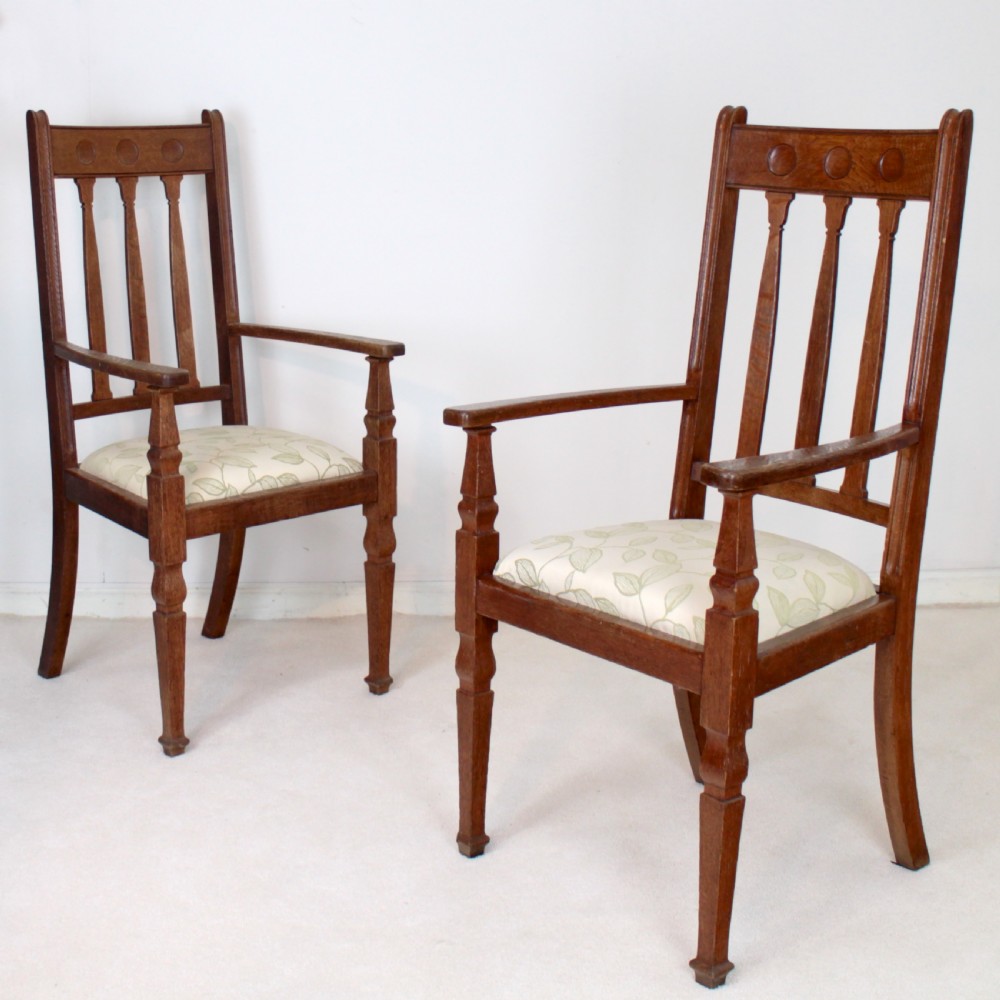 A pair of medium brown oak Arts and Crafts armchairs with carved circular back details, vertical splats, turned front legs, and light-coloured patterned seat cushions.