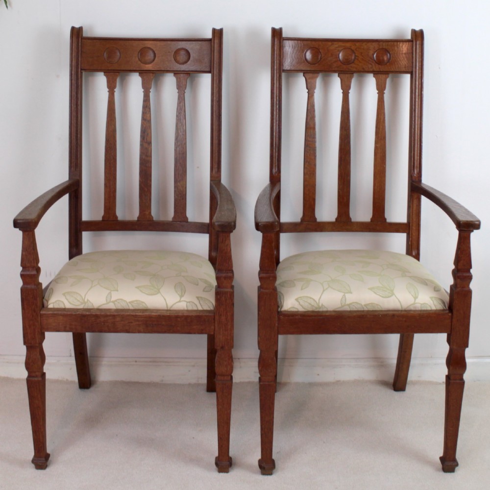 A pair of medium brown oak Arts and Crafts armchairs with carved circular back details, vertical splats, turned front legs, and light-coloured patterned seat cushions. View III