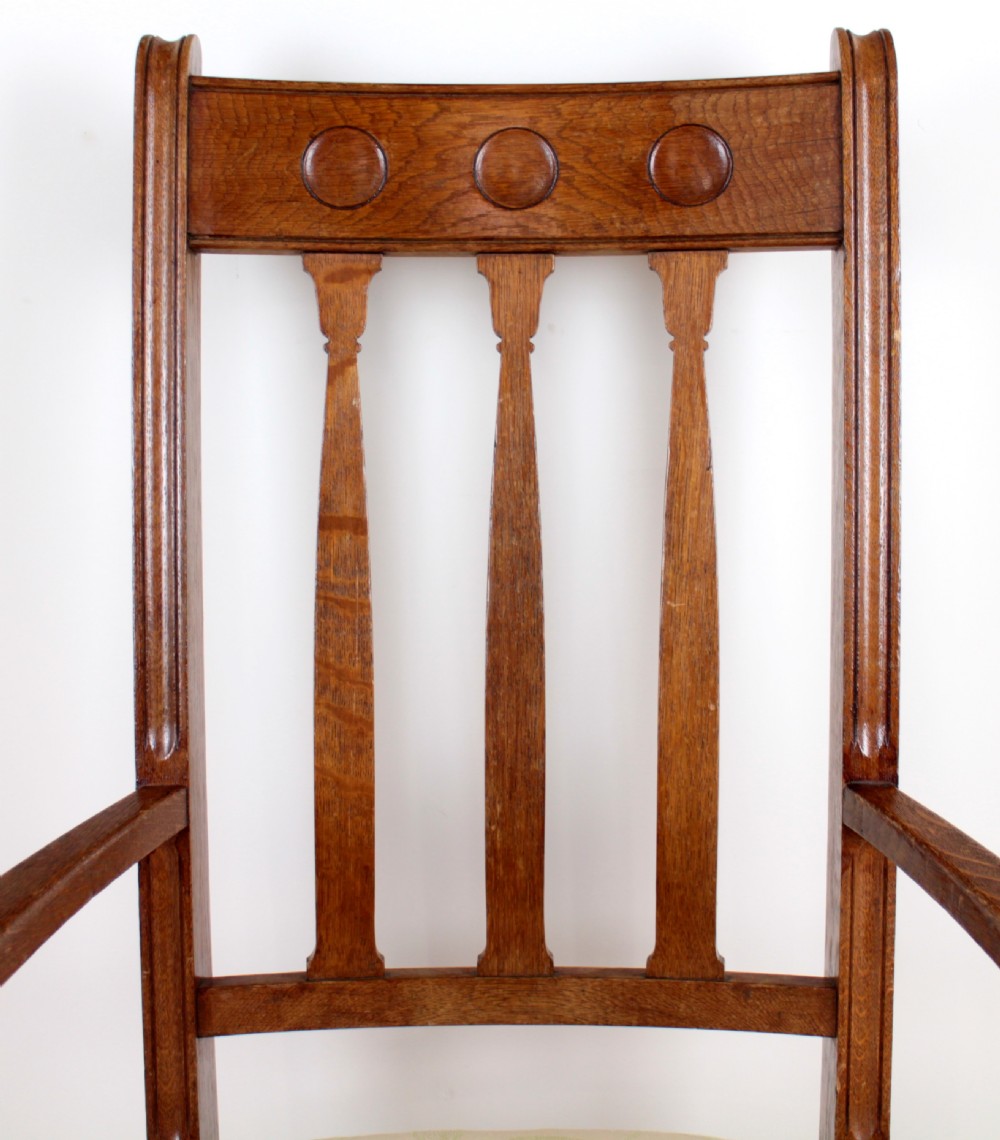 A pair of medium brown oak Arts and Crafts armchairs with carved circular back details, vertical splats, turned front legs, and light-coloured patterned seat cushions. View VI