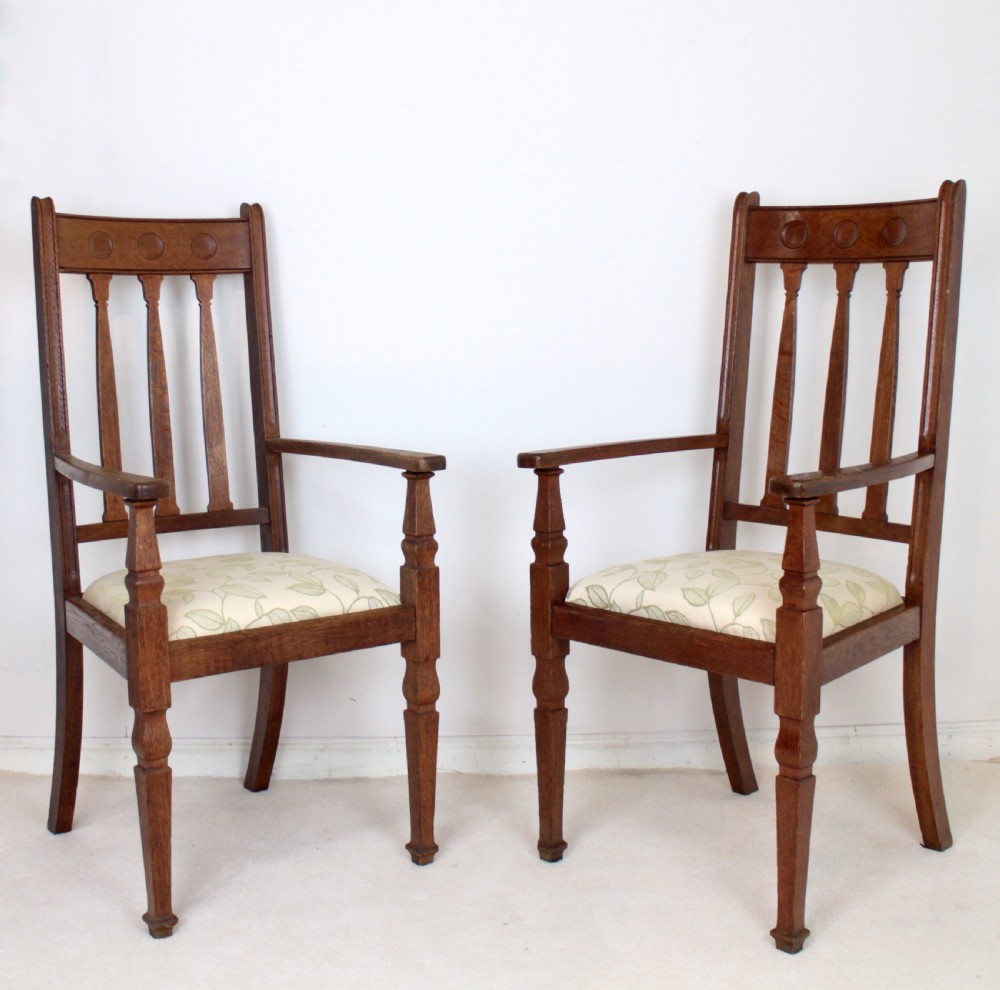 A pair of medium brown oak Arts and Crafts armchairs with carved circular back details, vertical splats, turned front legs, and light-coloured patterned seat cushions. View VII