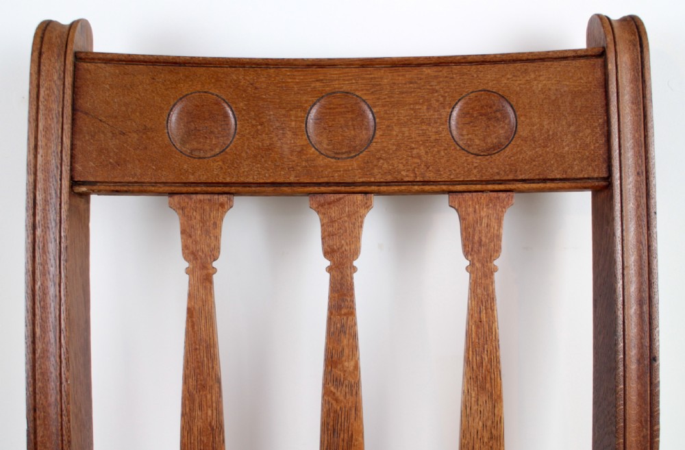 A pair of medium brown oak Arts and Crafts armchairs with carved circular back details, vertical splats, turned front legs, and light-coloured patterned seat cushions. View XV
