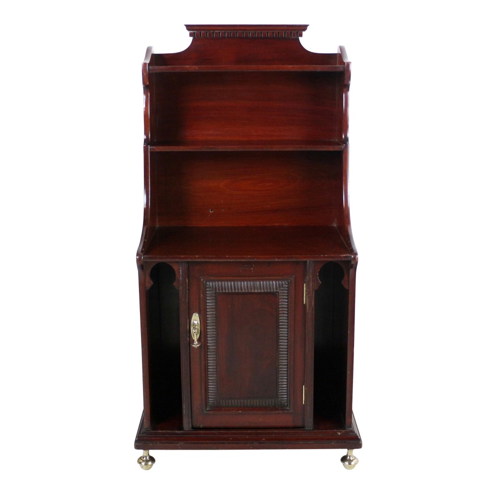 A dark mahogany Arts and Crafts waterfall bookcase featuring tiered open shelves above a single door cupboard with Moorish side cutouts and brass castors.