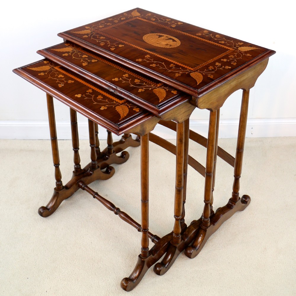 A set of three nested wooden side tables featuring detailed marquetry inlays of foliage and central vignettes, supported by slender turned legs and scroll feet.