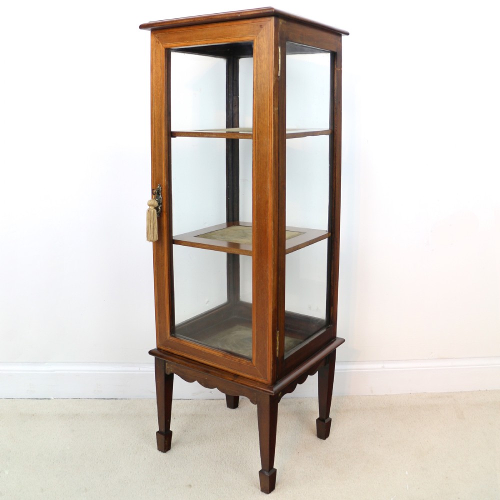 A tall Edwardian mahogany glazed bijouterie cabinet featuring glass panels on all sides, two interior shelves, square tapered legs, and satinwood crossbanding.