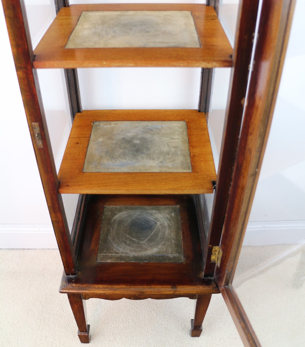 A tall Edwardian mahogany glazed bijouterie cabinet featuring glass panels on all sides, two interior shelves, square tapered legs, and satinwood crossbanding. View XXI