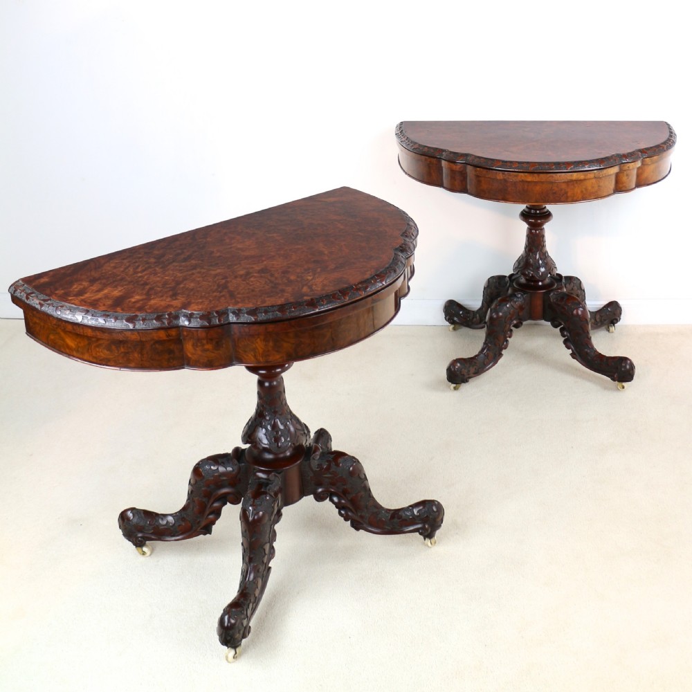 A pair of Victorian burr walnut fold-over card tables featuring shaped demi-lune tops, carved aprons, central turned pedestals, and carved quadform legs with castors.