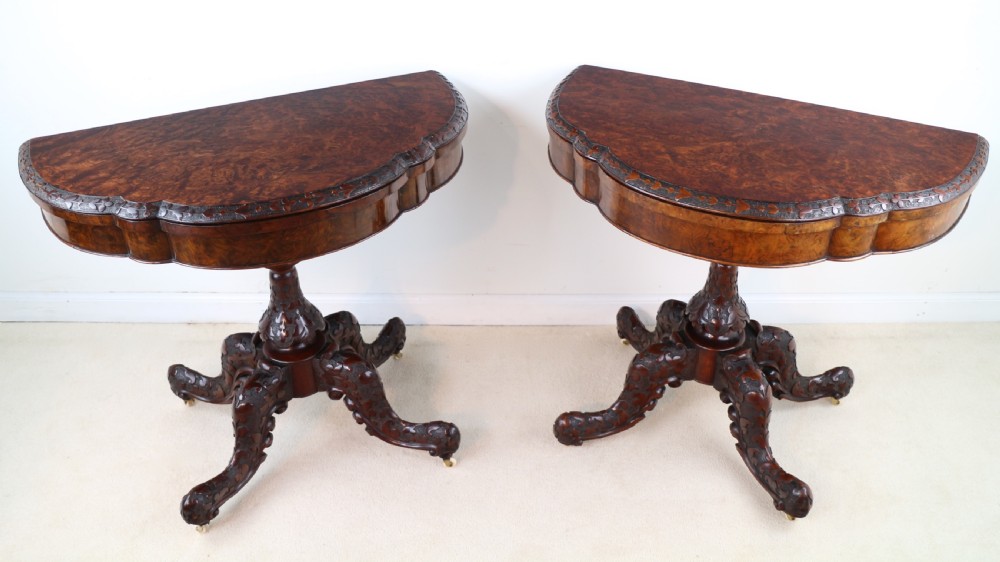 A pair of Victorian burr walnut fold-over card tables featuring shaped demi-lune tops, carved aprons, central turned pedestals, and carved quadform legs with castors. View V