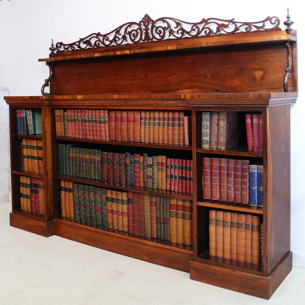 An antique Regency open rosewood bookcase featuring an inverted breakfront profile, multi-tier shelving, and an intricately carved fretwork gallery.