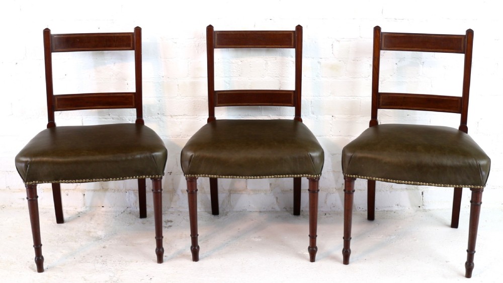 A set of twelve George III mahogany dining chairs featuring rectangular bar-back cresting rails with string inlay, green leather upholstered seats with stud detailing, and turned front legs. View VI