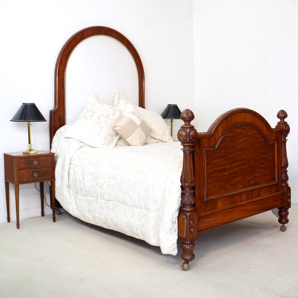 A Victorian mahogany double bed featuring a high arched upholstered headboard, a flame mahogany footboard with carved posts, and brass castors.