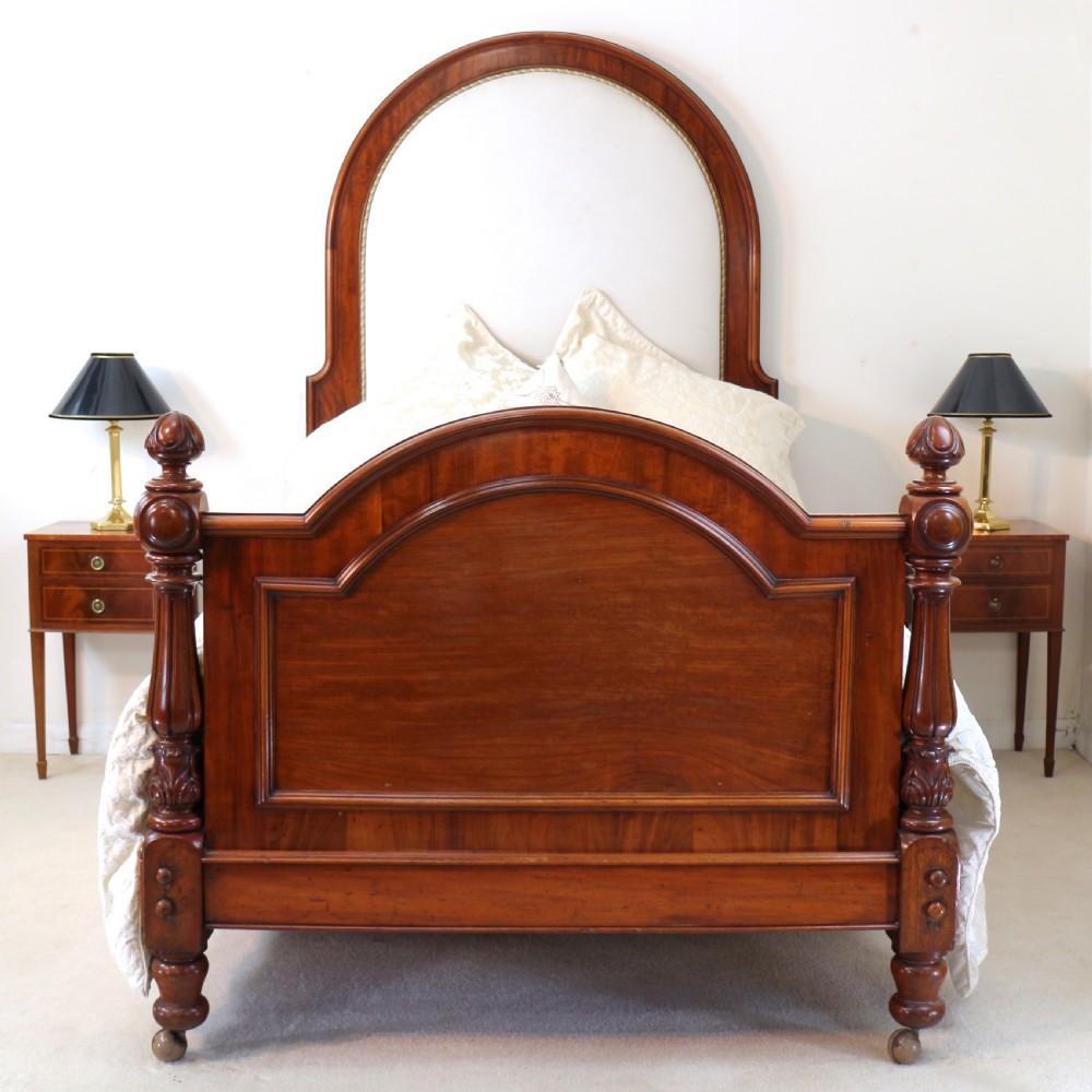 A Victorian mahogany double bed featuring a high arched upholstered headboard, a flame mahogany footboard with carved posts, and brass castors. View VI