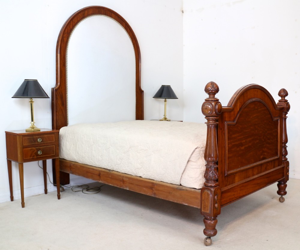 A Victorian mahogany double bed featuring a high arched upholstered headboard, a flame mahogany footboard with carved posts, and brass castors. View XV