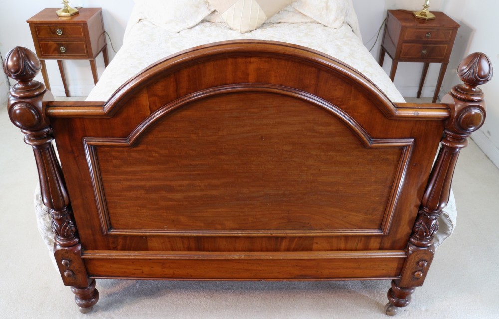 A Victorian mahogany double bed featuring a high arched upholstered headboard, a flame mahogany footboard with carved posts, and brass castors. View XVIII
