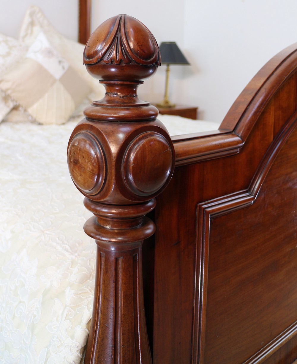A Victorian mahogany double bed featuring a high arched upholstered headboard, a flame mahogany footboard with carved posts, and brass castors. View XIX
