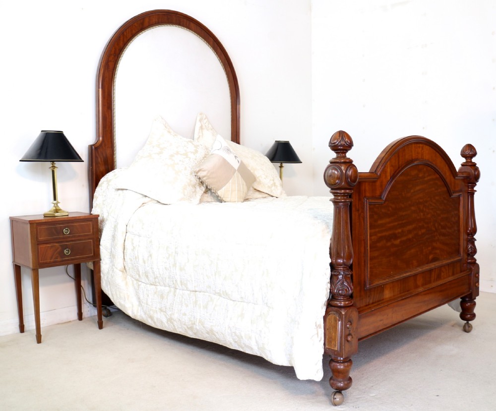 A Victorian mahogany double bed featuring a high arched upholstered headboard, a flame mahogany footboard with carved posts, and brass castors. View XX