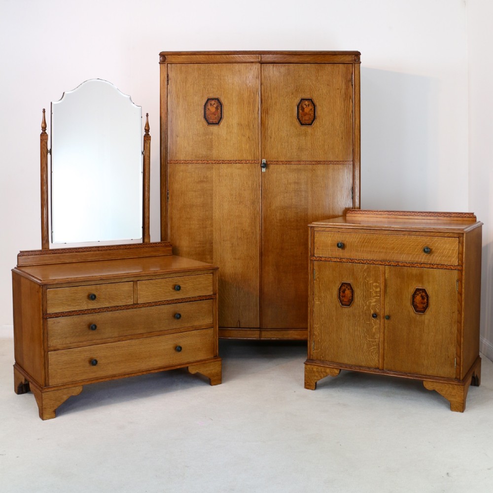 A three-piece golden oak Art Deco bedroom suite featuring a double wardrobe, dressing table with an arched mirror, and a small cupboard chest, all decorated with marquetry medallions.