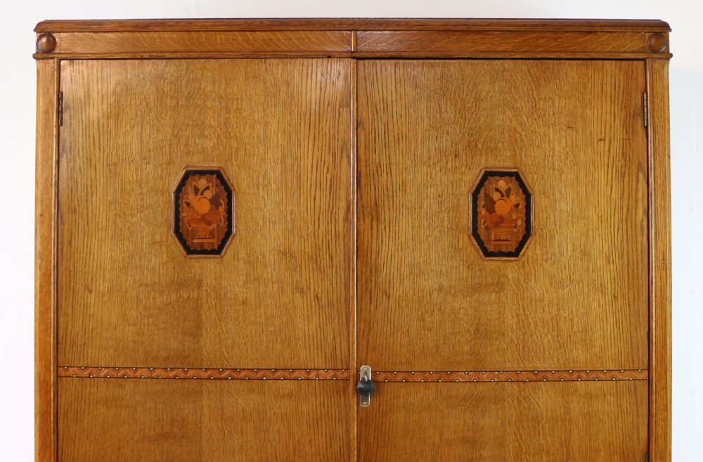 A three-piece golden oak Art Deco bedroom suite featuring a double wardrobe, dressing table with an arched mirror, and a small cupboard chest, all decorated with marquetry medallions. View V