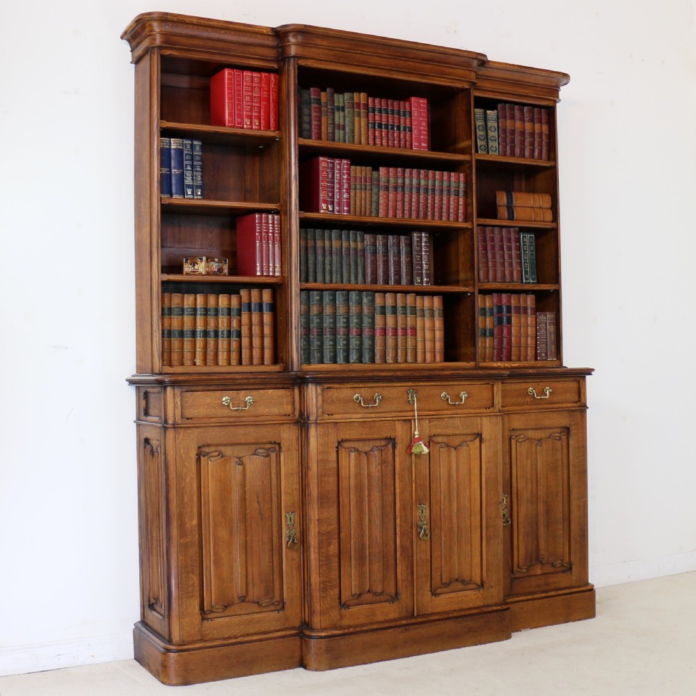 A large Victorian Gothic oak breakfront bookcase featuring open upper shelves filled with books and carved lower cupboard doors.
