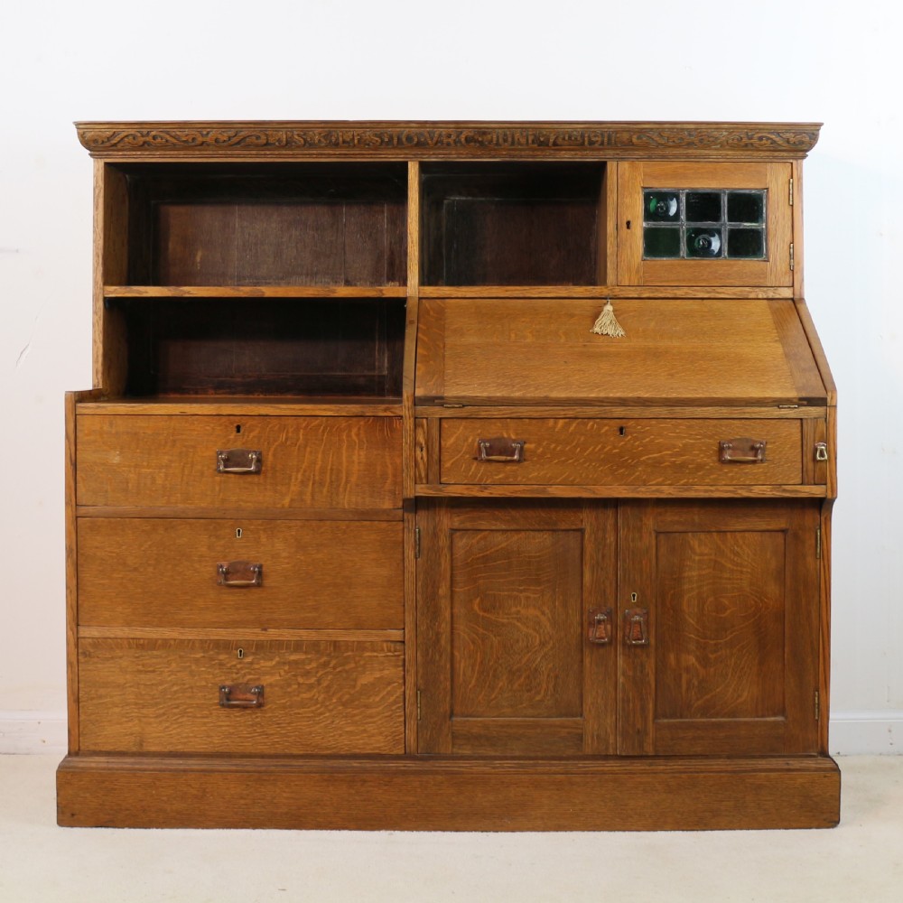 An Arts and Crafts quarter-sawn oak bureau bookcase featuring an asymmetrical design with open shelves, a fall-front writing slope, a leaded green glass cabinet, and drawers with copper handles.