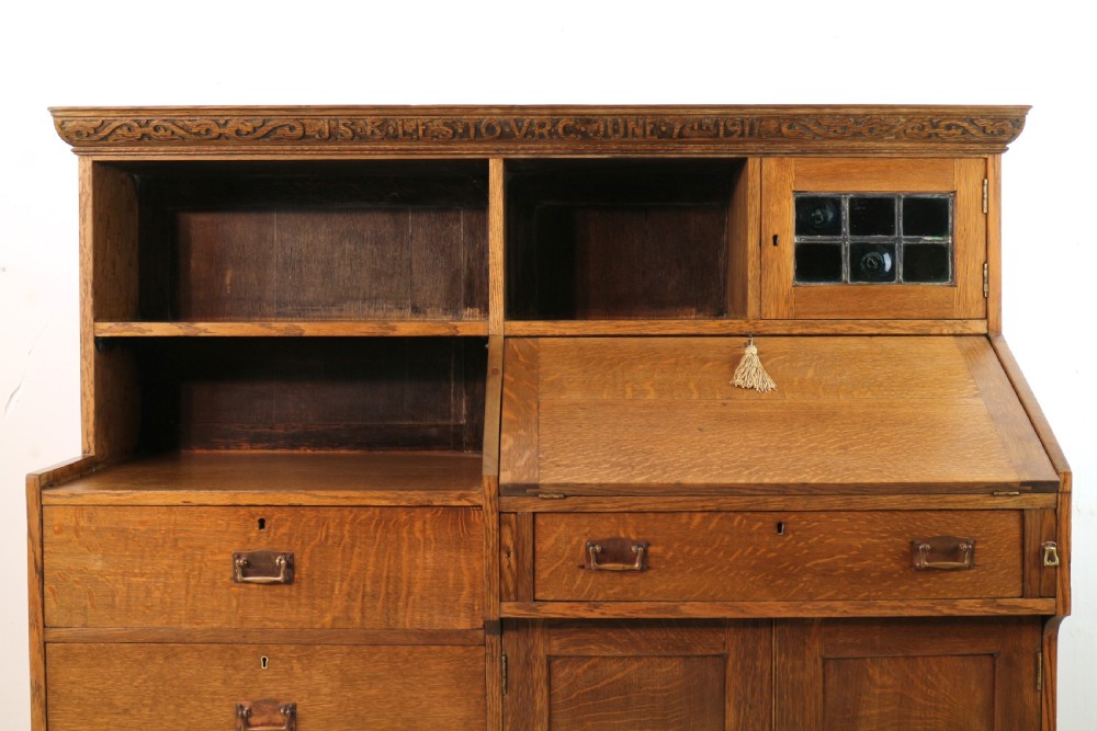 An Arts and Crafts quarter-sawn oak bureau bookcase featuring an asymmetrical design with open shelves, a fall-front writing slope, a leaded green glass cabinet, and drawers with copper handles. View II